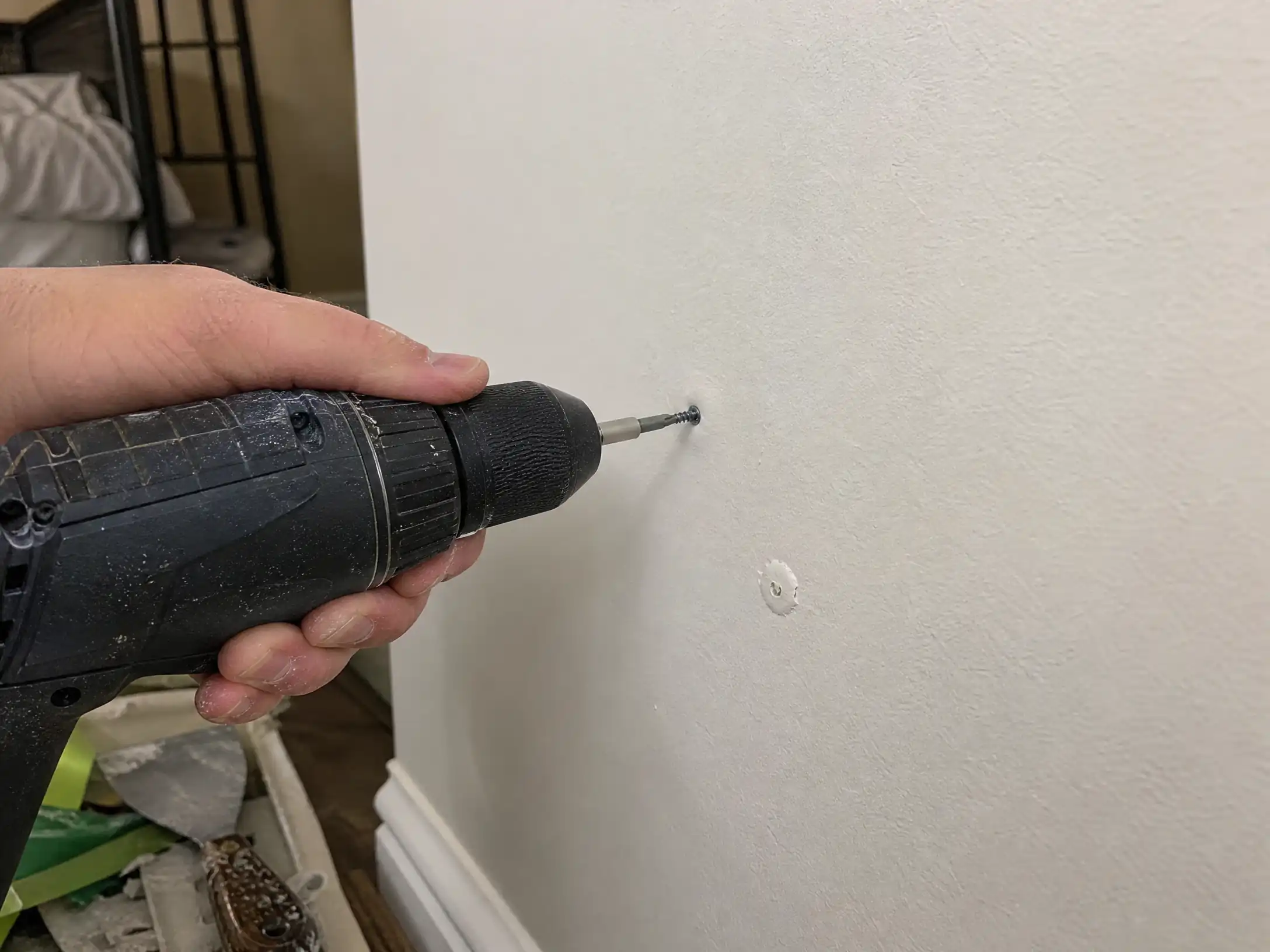 A cordless drill driving a drywall screw into a wall just above a nail pop, re-anchoring the panel