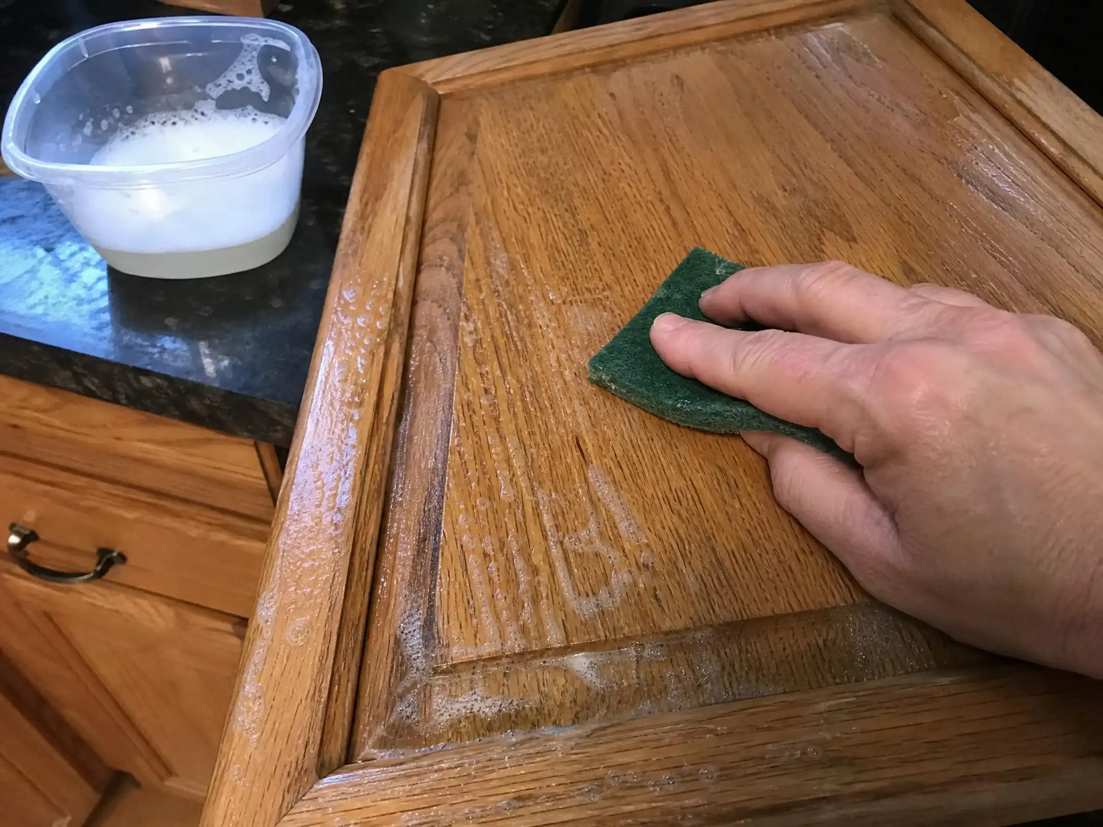 Homemade degreasing solution being scrubbed onto oak kitchen cabinet doors with a green scouring pad after six years of baked-on cooking grease